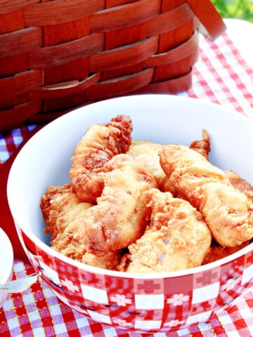 A red and white checked bowl filled with Waffle Batter Fried Chicken Tenders along with a picnic basket.