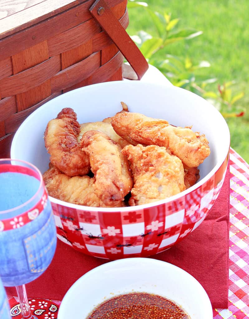 A vertical photo of a red and white checked bowl filled with Waffle Batter Fried Chicken tenders with a picnic basket in the background along with a glass of wine.