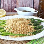 A white platter filled with Asparagus with Breadcrumbs along with a fresh lemon in the background and salt and pepper containers.