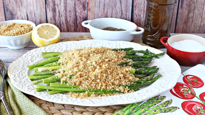 A white platter filled with Asparagus with Breadcrumbs along with a fresh lemon in the background and salt and pepper containers.