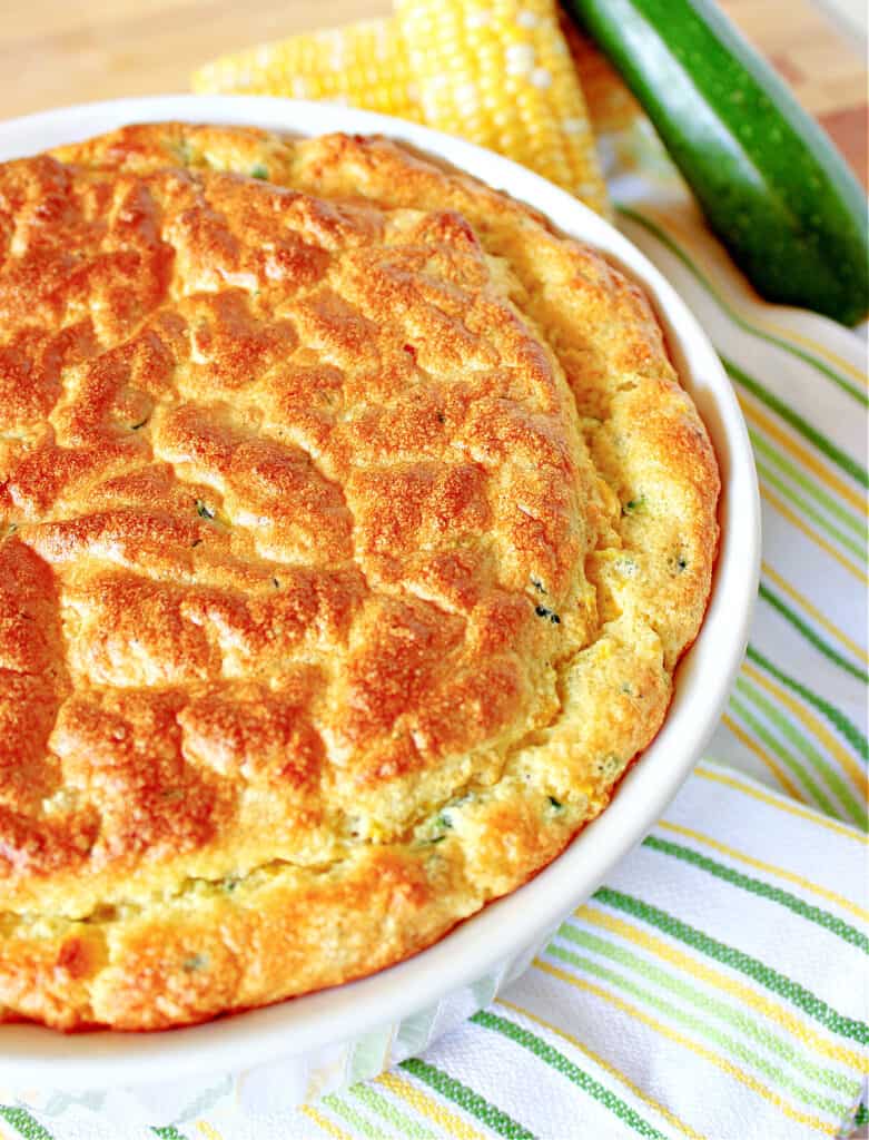 A vertical closeup of a Zucchini and Sweet Corn Soufflé in a round white casserole dish with a green and white stripped napkin on the side.