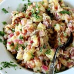 German Potato Salad in a white bowl along with a spoon and a garnish of parsley.