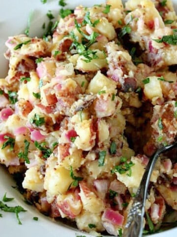 German Potato Salad in a white bowl along with a spoon and a garnish of parsley.