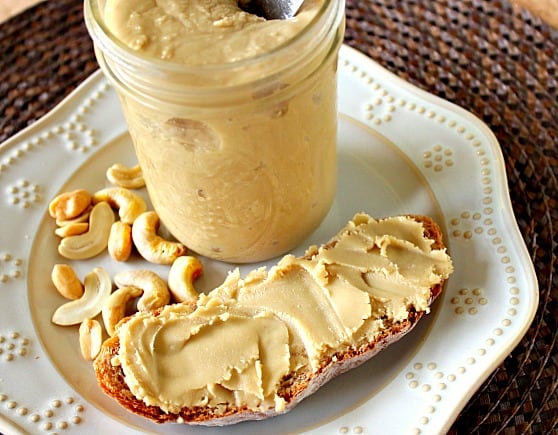 A slice of bread with spread with cashew peanut butter on a white plate with a jar.