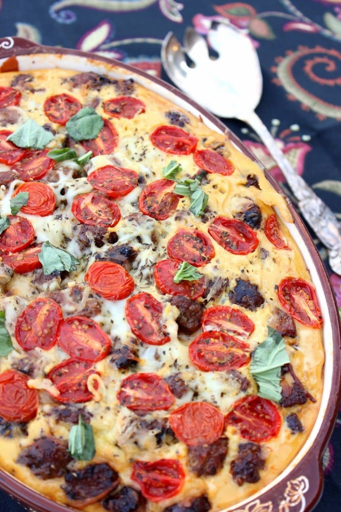 A colorful Lasagna Strata in a casserole dish with fresh basil, tomatoes, and sausage.