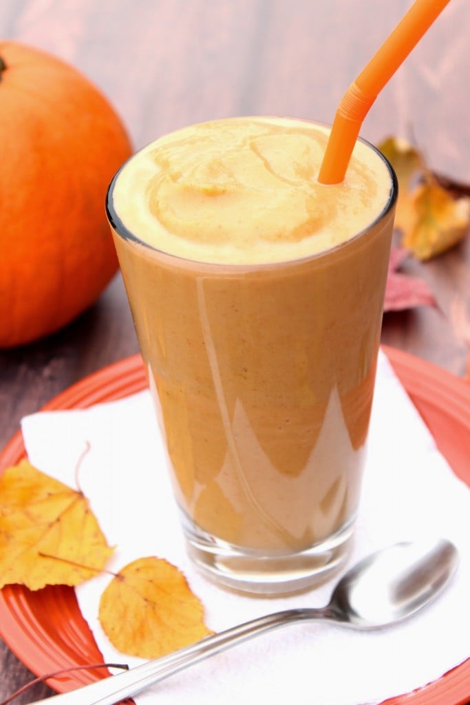A tall glass filled with a Low Calorie Pumpkin Pie Milkshake and an orange straw and a pumpkin in the background.