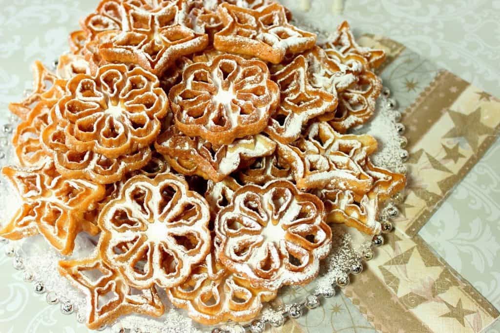 A horizontal photo of a plate of fried rosette cookies on a glass plate with powder sugar, and star napkins.