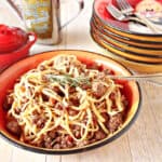 A bowl filled with Linguine with Lamb along with a fork and a stack of bowls in the background.