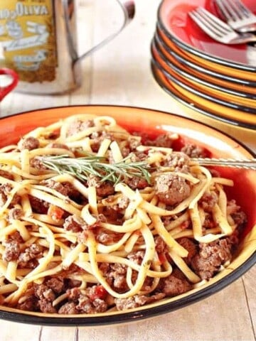 A bowl filled with Linguine with Lamb along with a fork and a stack of bowls in the background.