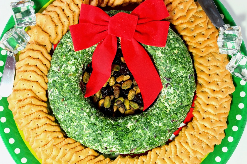 Overhead closeup photo of a goat cheese Christmas wreath appetizer on a round platter with crackers and a red bow.