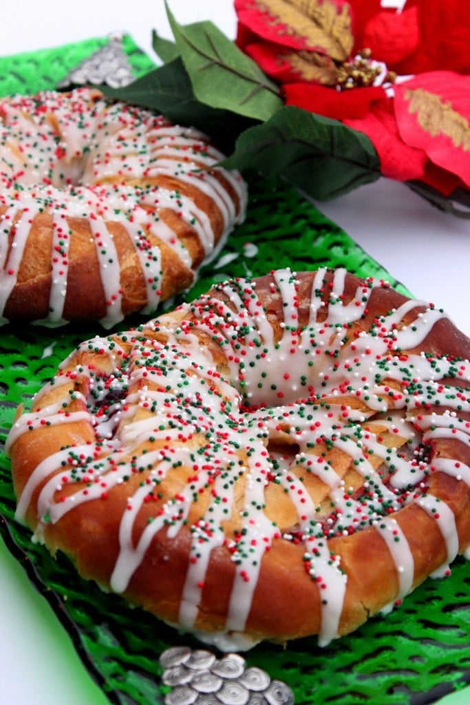 A vertical image of two cranberry Kringles on a green Christmas platter with icing and Christmas sprinkles.