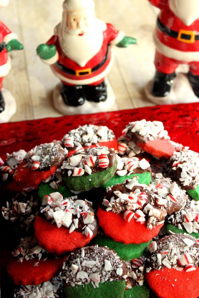 A vertical closeup photo of red and green cookies dipped in chocolate with crushed peppermint candy with fun Santa figures in the background.