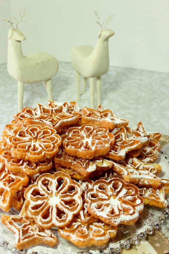 A vertical photo of a glass plate loaded with fried rosette snowflake cookies with confectioner sugar dusting.