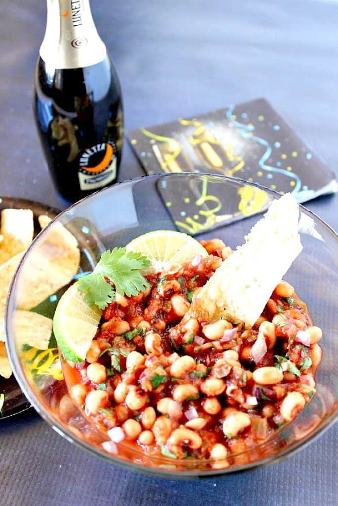 Black-Eyed Pea Salsa in a glass bowl with a bottle of champagne in the background along with a napkin.