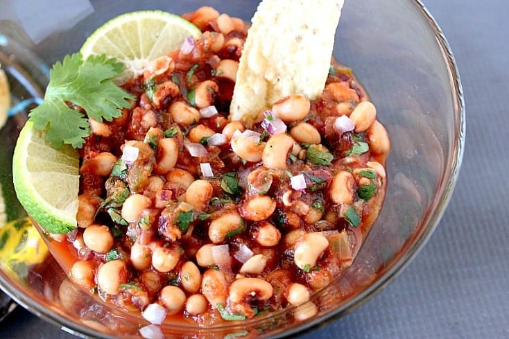 An offset horizontal image of a glass bowl filled with Black-Eyed Pea Salsa along with lime wedges and fresh cilantro.