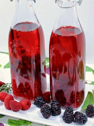 Two tall bottles filled with Homemade Berry Infused Vinegar on a white plate with fresh berries and herbs.