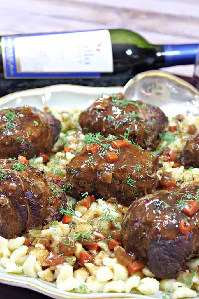 A vertical closeup of a platter of German Beef Rouladen with spaetzle and a bottle of wine in the background.