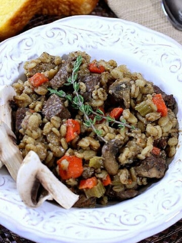 A pretty white bowl filled with Beef Barley and Mushroom Stew with fresh rosemary on top.