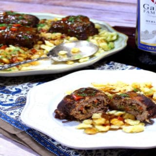 A plate of German beef rouladen in the foreground and a platter of German beef rouladen in the background with a bottle of wine