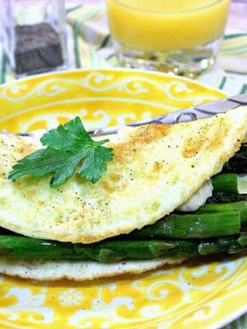 An Egg White Omelet with Asparagus spears on a yellow plate with a glass of orange juice in the background.