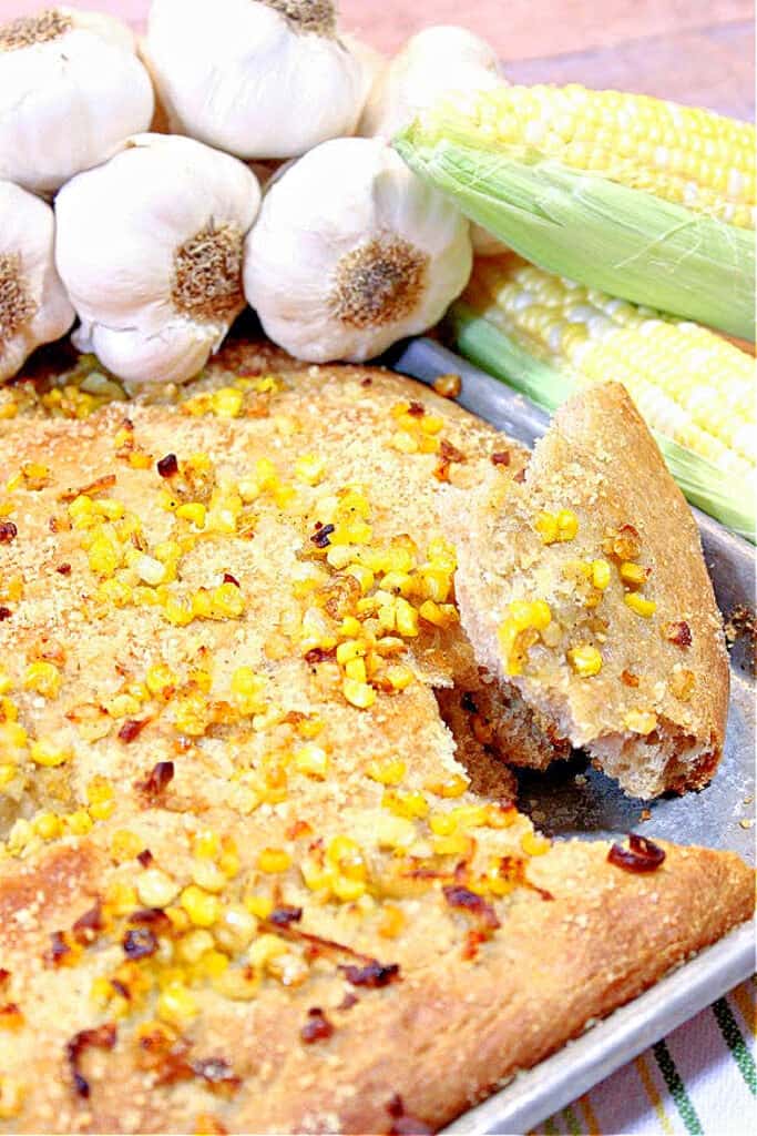 A vertical closeup of a Garlic Focaccia with Sweet Corn on a sheet tray topped with corn, shallots with garlic and corn in the background.