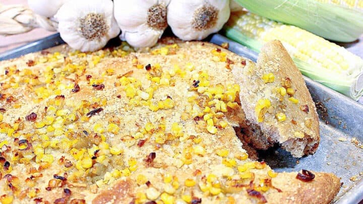 A Garlic Focaccia with Sweet Corn on a sheet tray along with garlic bulbs and fresh corn in the background.