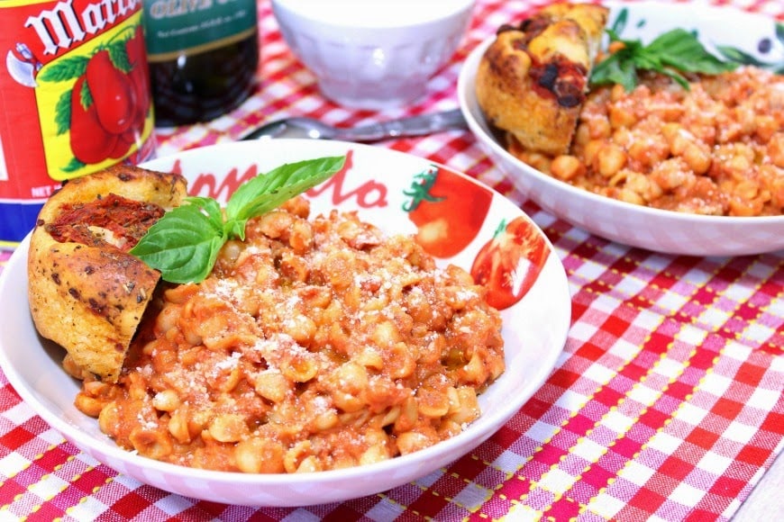 A horizontal photo of two bowls of tomato bacon pasta on a red and white check table cloth with fresh basil leaves. Valentine's day dinner recipe roundup.