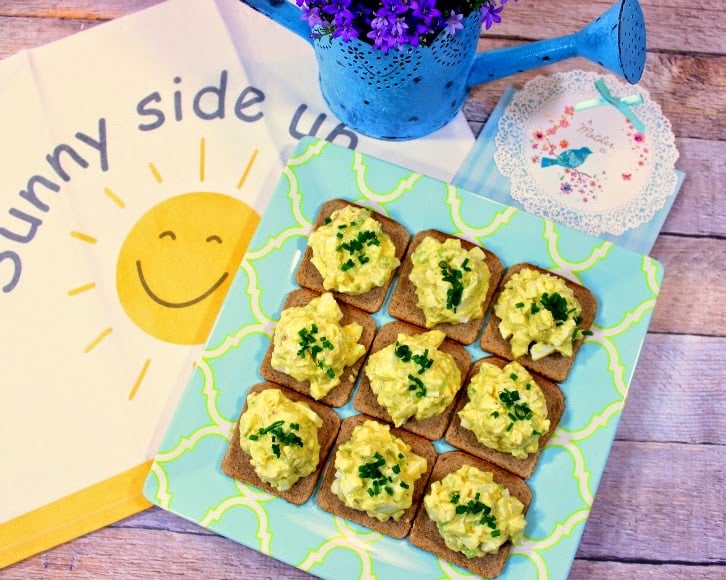 Overhead picture of a square plate of egg salad appetizers with a smiling face sunshine napkin and a blue watering can with purple flowers.