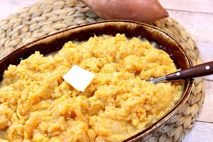 A vertical photo of a brown bowl filled with cauliflower sweet potato mashers, butter, and a spoon.