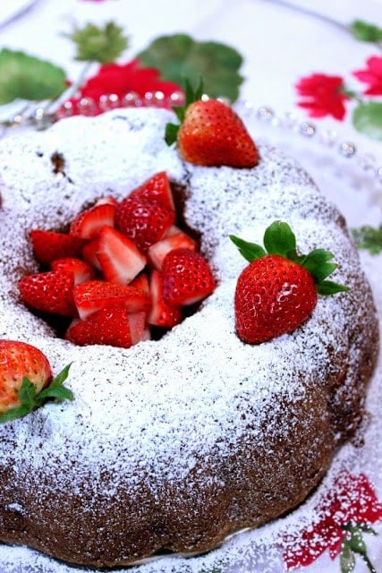 Old-Fashioned Strawberry Rhubarb Bundt Cake Vertical closeup photo of a bundt cake on a glass plate with fresh strawberries on top and a confectioners sugar dusting.