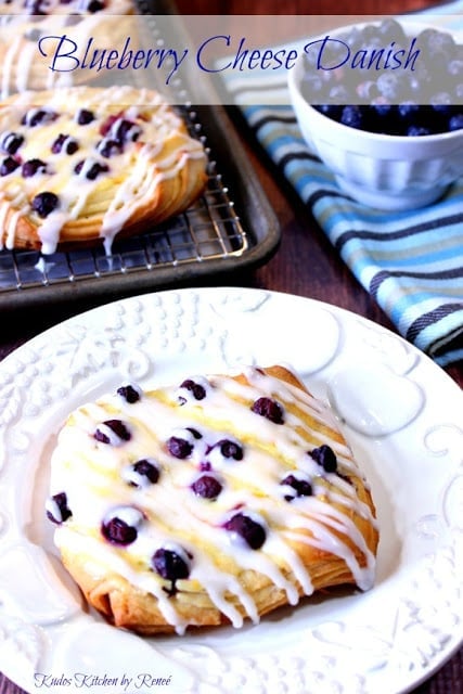 A blueberry danish on a white plate with more in the background on a baking sheet.