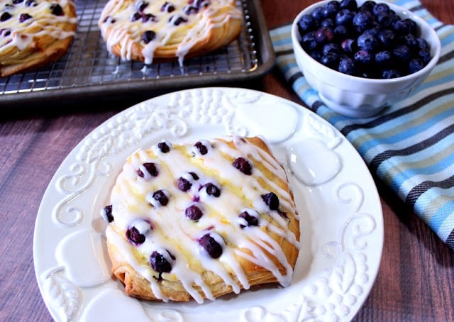 Blueberry cheese danish on a white plate with a bowl of fresh blueberries in the background.