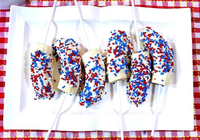 An overhead photo of white chocolate frozen bananas on a plate with red, white, and blue sprinkles.