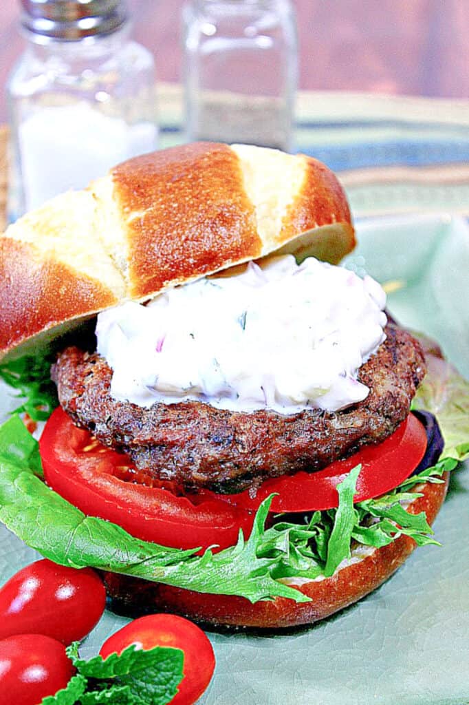 A colorful vertical image of a Lamb Burger with Feta and Tzatziki on a pretzel bun along with lettuce and tomatoes.