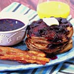 A stack of Whole Wheat Blueberry Pancakes on a blue platter with bacon and a bowl of Blueberry Maple Syrup on the side.