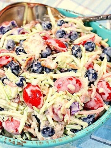 A blue bowl filled with Broccoli Blueberry Slaw with Bacon along with a large serving spoon.