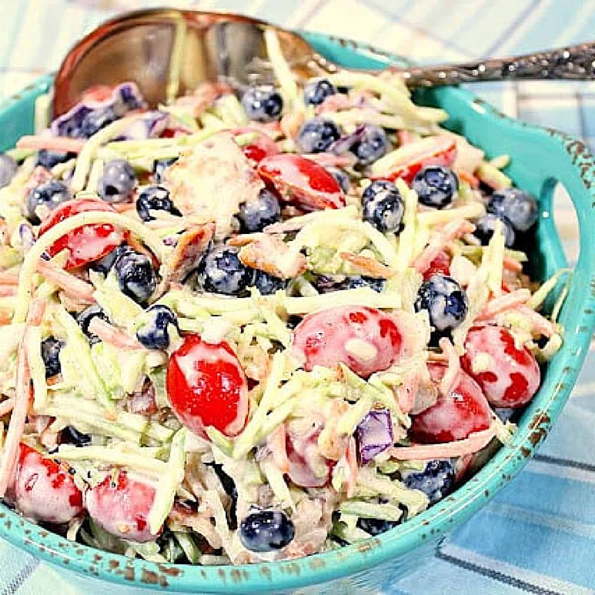A blue bowl filled with Broccoli Blueberry Slaw with Bacon along with a large serving spoon.