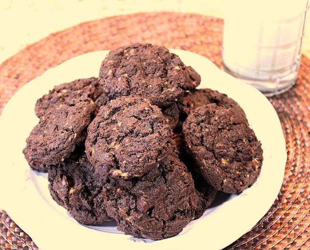 A stacked bunch of Chocolate Oat Toffee Chip Cookies on a plate with a glass of milk in the background.