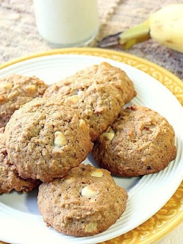 A yellow and white plate filled with Banana Walnut Cookies with a glass of milk and a banana in the background.