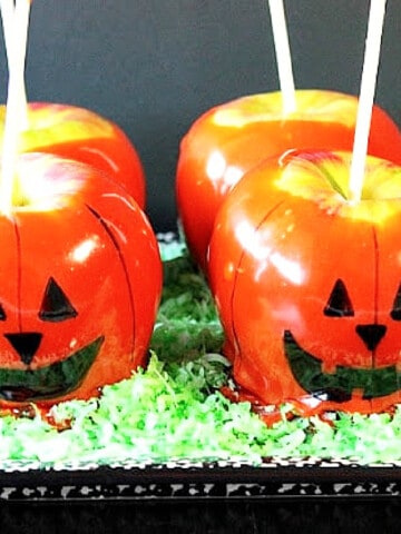 Four Jack-O-Lantern Caramel Apples on a platter with green colored coconut.