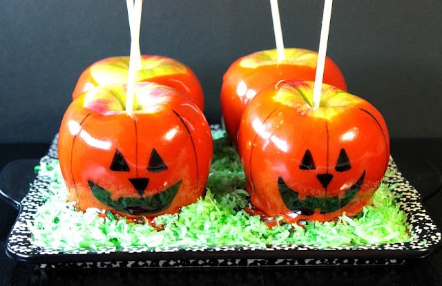 Four Jack-O-Lantern Caramel Apples on a platter with green colored coconut.