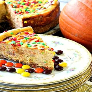 A slice of Reese's Pieces Pumpkin Cheesecake on a stack of china plate with the entire cheesecake in the background.