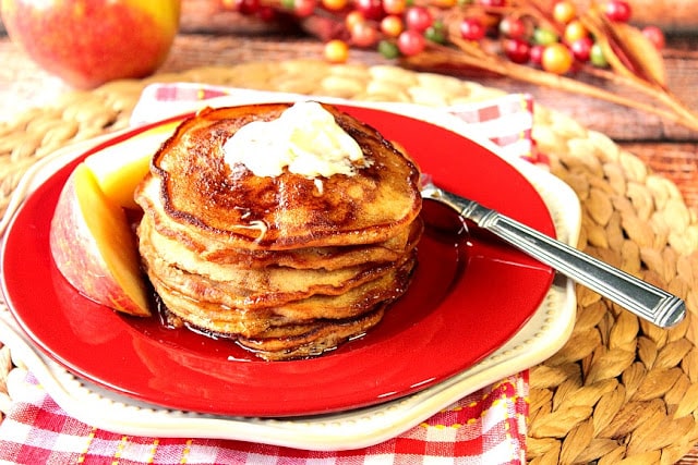 A stack of Apple Cider Pancakes with apple slices on a red plate with butter.