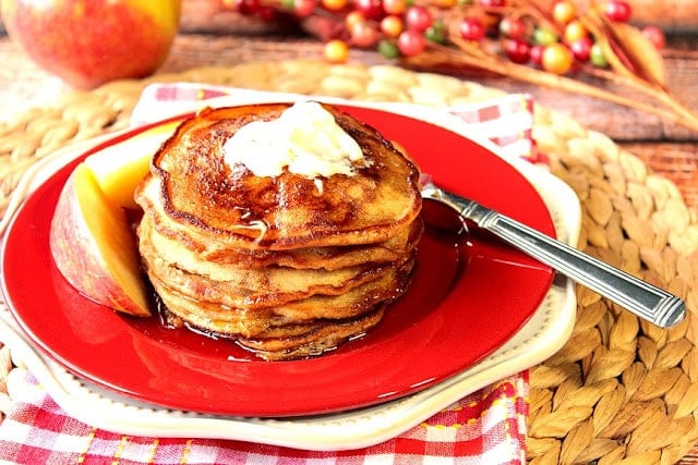 A stack of Apple Cider Pancakes with apple slices on a red plate with butter.