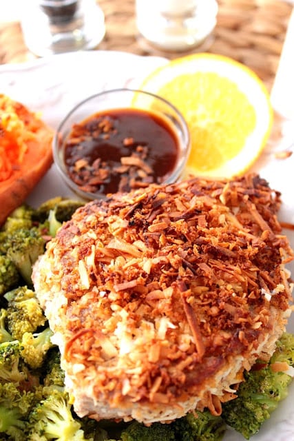 Vertical photo of browned coconut crusted tuna steak with an orange slice, dipping sauce, and broccoli on a white plate.