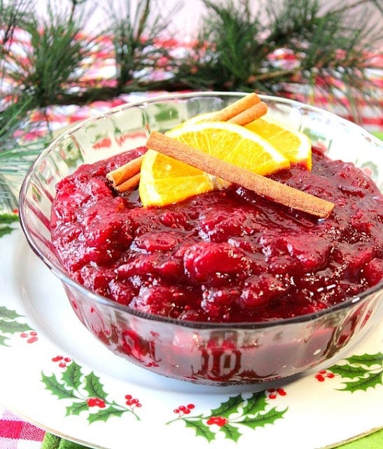 A vertical photo of a glass bowl with cranberry sauce, orange slices, and cinnamon sticks.