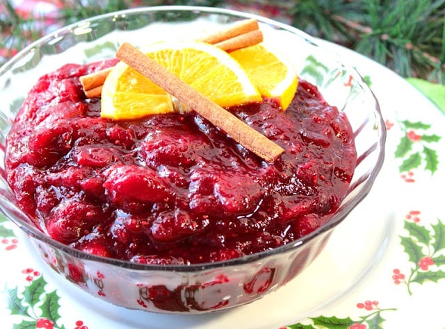 Closeup horizontal photo of a glass bowl filled with pineapple cardamom cranberry sauce with cinnamon stick and orange wedges on top. Christmas dinner recipe roundup.