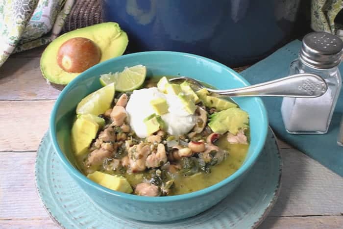 A blue bowl of black-eyed pea chicken chili with avocado and sour cream and a soup spoon.