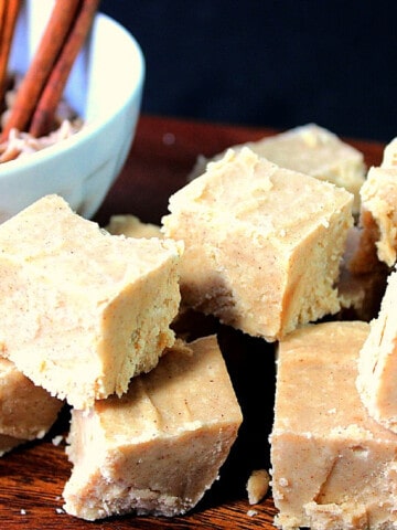 A stack of Cinnamon Fudge in the foreground and a bowl of cinnamon chips and sticks in the background.