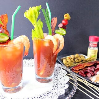 Two tall cocktail glasses filled with Bacon Bloody Mary along with shrimp, celery, scallions, and sausage.